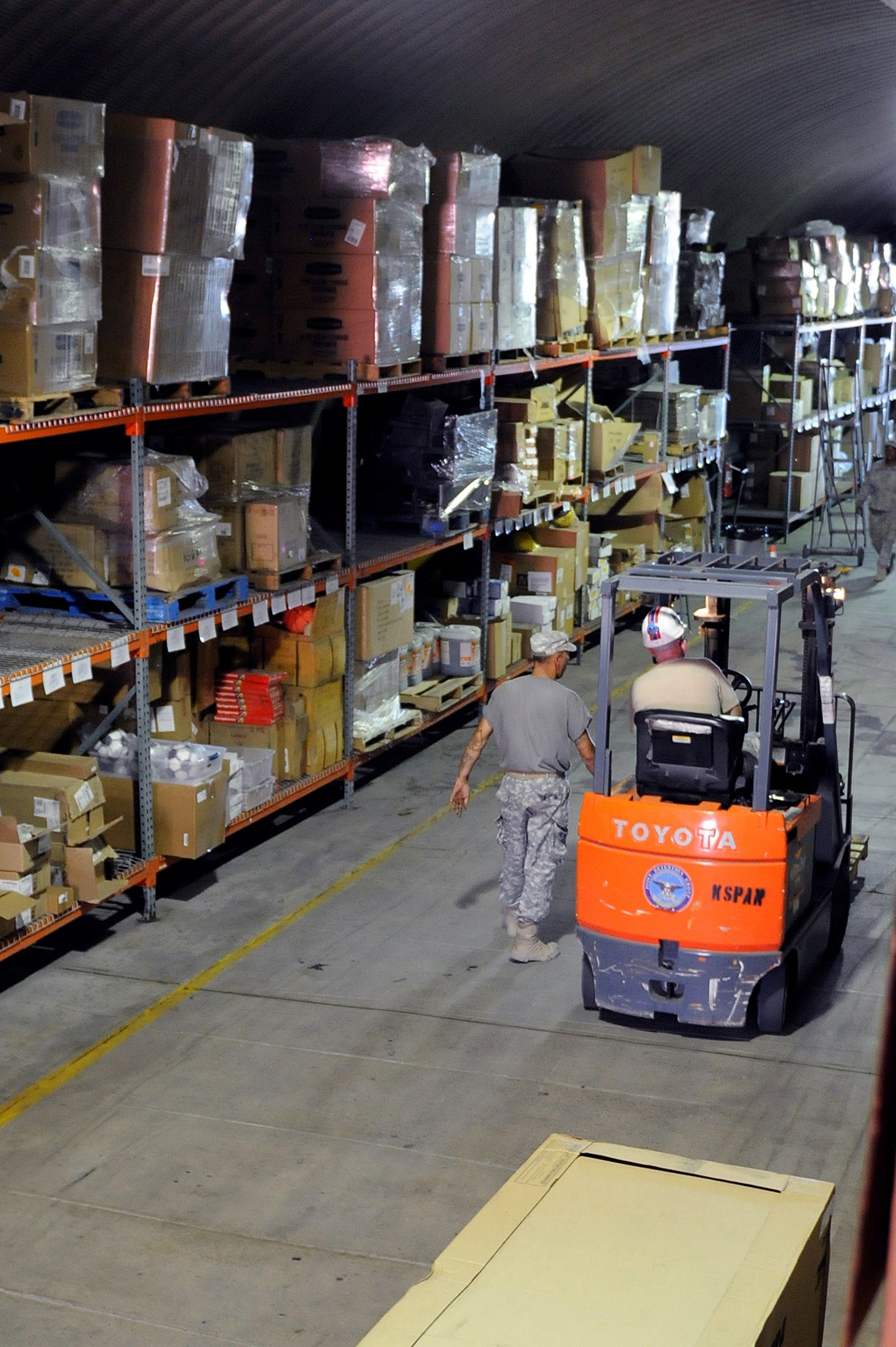 Ein Mann bedient einen Gabelstapler in einem Lagerhaus mit Stapeln von Kartons und anderen Gegenständen auf Regalen, mit einem einzelnen Karton am Boden des Bildes.