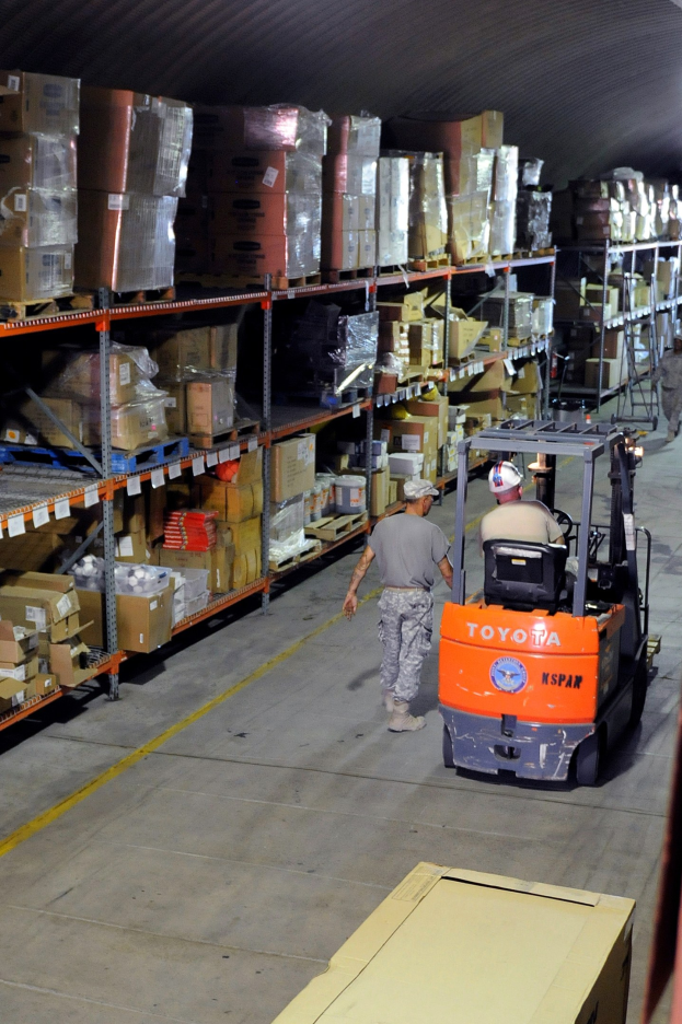 Ein Mann bedient einen Gabelstapler in einem Lagerhaus mit Stapeln von Kartons und anderen Gegenständen auf Regalen, mit einem einzelnen Karton am Boden des Bildes.