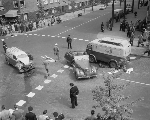 Eine schwarz-weiße Szenerie eines Autounfalls an einer Straßenkreuzung, mit mehreren Fahrzeugen einschließlich eines Lieferwagens, einer Gruppe von Menschen drumherum und Gebäuden mit Fenstern, Bäumen, Pfählen und Texttafeln im Hintergrund.