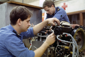 Zwei Männer in blauen Uniformen arbeiten an einem Flugzeugtriebwerk in einer Fabrik, mit einem roten Kreuzsymbol an der Wand im Hintergrund.