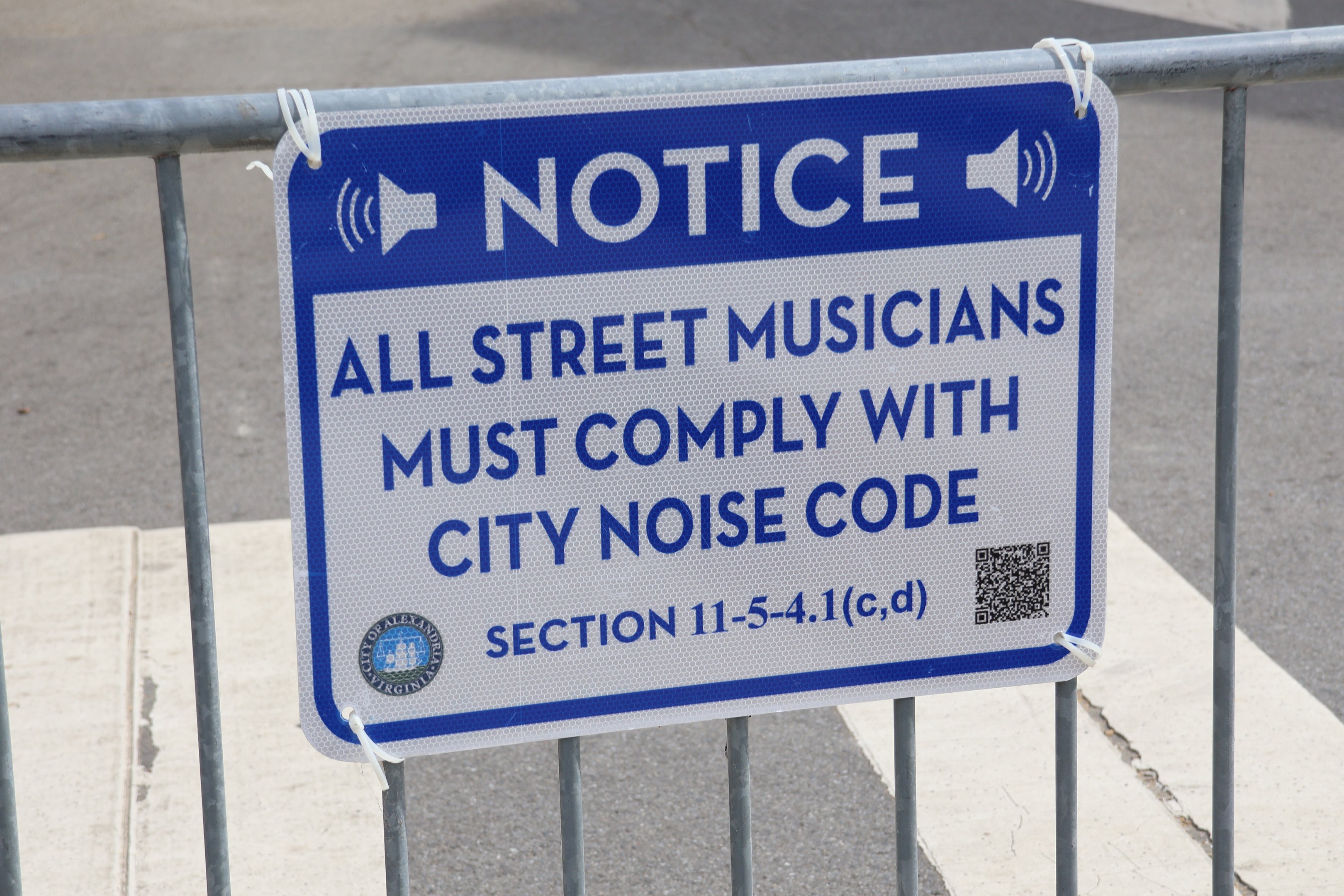 Anschlagtafel an einem Zaun mit einem Schild, auf dem "Hinweis: Alle Straßenmusiker müssen die städtischen Lärmvorschriften einhalten" steht und eine Straße im Hintergrund.