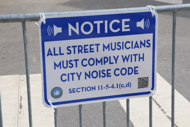 Anschlagtafel an einem Zaun mit einem Schild, auf dem "Hinweis: Alle Straßenmusiker müssen die städtischen Lärmvorschriften einhalten" steht und eine Straße im Hintergrund.