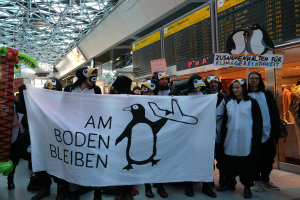 Gruppe von Menschen in Pinguinkostümen mit einem Banner vor einer Wand mit Pinguin-Plakaten, Schaufensterpuppen in Kleidern und anderen Gegenständen im Hintergrund.