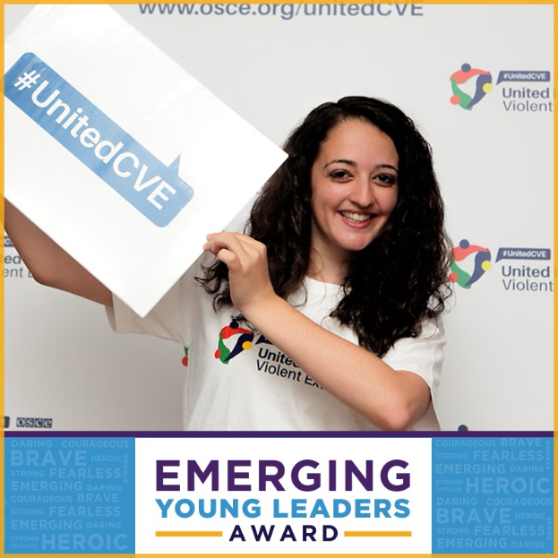 Eine strahlende Frau in einem weißen T-Shirt und Pferdeschwanz hält ein Schild mit der Aufschrift "Emerging Young Leaders Award" hoch, das Freude und Erfolg zeigt.