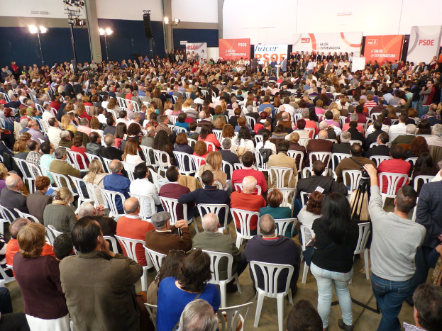 Ein volles Stadion mit vielen sitzenden und stehenden Menschen, eine Bühne mit zwei Rednern auf einem Podium und mehrere Plakate und Lichter in der Nähe.