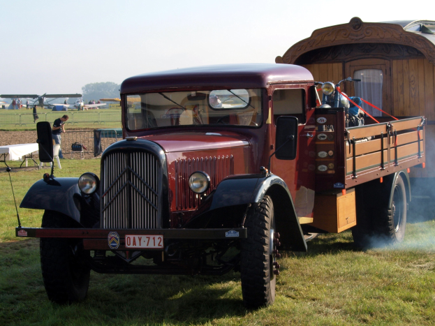 Ein roter Lkw neben einem Anhänger auf einer grünen Wiese mit Menschen in der Nähe, Flugzeuge im Hintergrund, Bäume, einen klaren blauen Himmel und Rauch, der aus dem Anhänger aufsteigt.