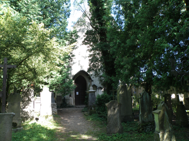 Friedhof mit zahlreichen Grabsteinen, Bäumen im Hintergrund, einem Gebäude mit einer Tür und einem klaren blauen Himmel.