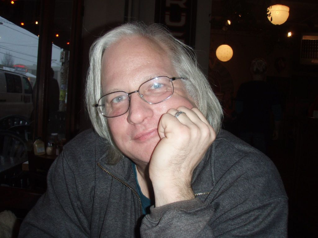 Ein Mann in der Mitte mit seiner Hand auf dem Gesicht, ein Tisch mit Gläsern und Flaschen links, ein Glas mit einem Van auf der Straße neben dem Tisch und Lichter rechts.