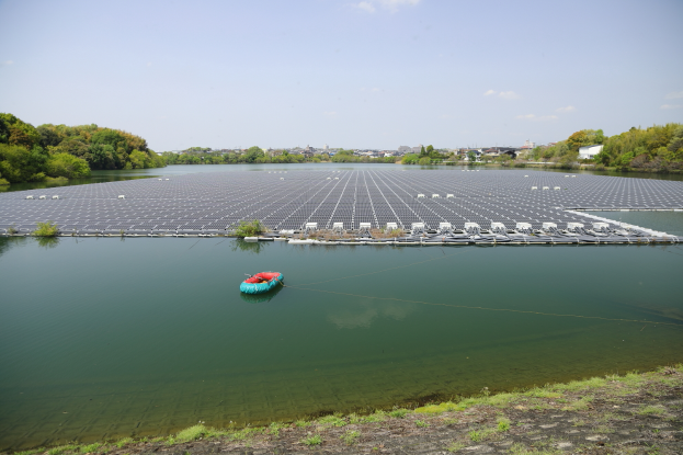 Ein kleines Boot treibt auf einem Gewässer, das von grünem Gras und Bäumen umgeben ist, mit Gebäuden und einem klaren blauen Himmel im Hintergrund und Solarpanelen auf der Wasseroberfläche.