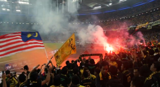 Große Menschenmenge in einem Stadion mit Feuerwerk, Rauch und Fahnen, darunter eine Person mit einer Kamera, sowie Schilder mit der Aufschrift "Malaysia vs Malaysia Live-Streaming".