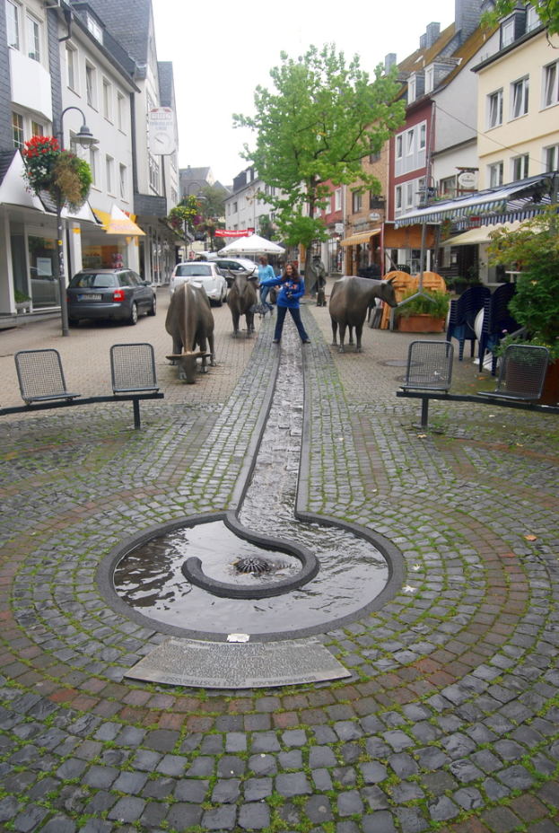 Rinder und Menschen auf einer Straße mit einem Brunnen, Bänken, Kraftfahrzeugen, Sonnenschirmen, Bäumen, Gebäuden, Straßenlaternen und Wolken am Himmel.