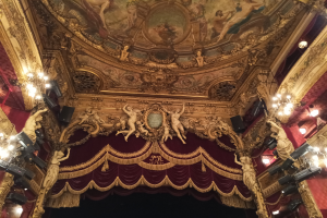 Innere eines Theaters mit einem roten Vorhang, Kronleuchtern, Skulpturen, Lichtern und einer bemalten Decke, mit einer Bühne und ein paar Menschen unten.