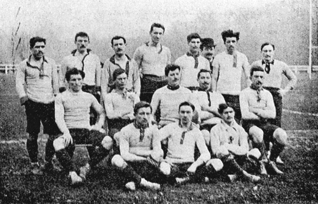 Ein Schwarz-Weiß-Foto einer Fußballmannschaft aus den 1920er Jahren, mit einigen stehenden und anderen sitzenden Spielern, sowie Pfählen, Bäumen und einem klaren Himmel im Hintergrund.
