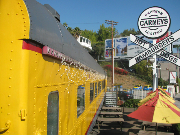 Eine mobile Kantine ist in der Nähe von Bänken und Treppen geparkt, mit Schirmen, Pflanzen und einem Geländer in der Nähe. Fahrzeuge, ein Mast, Schilder und eine Absperrung sind im Hintergrund zu sehen, sowie ein Gebäude, Bäume und der Himmel.