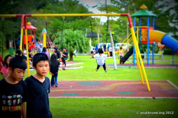 Kinder beim Spielen auf Geräten in einem Park mit Bäumen im Hintergrund.