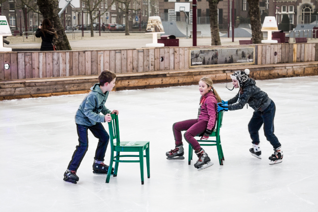 Kinder skifahren im Vordergrund eines Spielplatzes, mit drei Kindern und zwei Stühlen in der Mitte und Gebäuden, Bäumen, Bänken, Pfosten und einem Basketballfeld im Hintergrund.