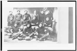 Ein Schwarz-Weiß-Foto einer Fussballmannschaft, mit einigen Spielern sitzend und einigen stehend, und Bällen auf dem Boden verstreut, mit Text unten.