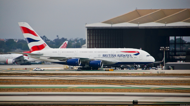 Ein Flugzeug befindet sich auf der Startbahn mit Straßenlaternen auf der rechten Seite, einem Auto in der Nähe und einem Schuppen im Hintergrund. In der Ferne sind Bäume, Gebäude und ein weiteres Flugzeug zu sehen, das auf einem offenen Gebiet geparkt ist, mit einem von Wolken erfüllten Himmel darüber.