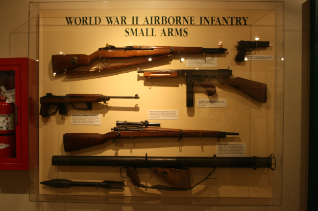 Vitrine mit verschiedenen Gewehren, ordentlich angeordnet, mit einem Feuerlöscher links und einer Wand im Hintergrund.