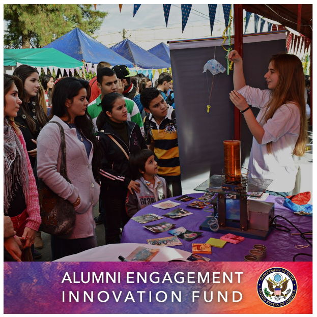 Eine Gruppe von Menschen steht um einen Tisch mit der Aufschrift "Alumni Engagement Innovation Fund", verschiedene Gegenstände auf dem Tisch, Zelte und Bäume im Hintergrund unter einem klaren blauen Himmel, mit Text und Logo unten auf dem Bild.