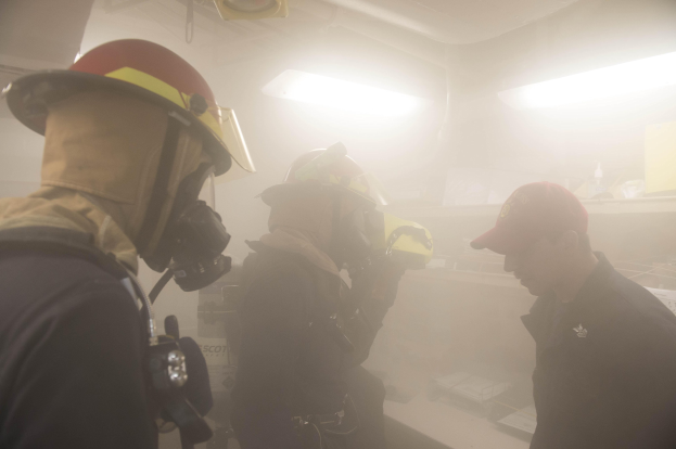 Feuerwehrleute in Helmen und Ausrüstung atmen Rauch während einer Übung aus, einer hält eine Kamera, mit Lampen und Equipment im Hintergrund.