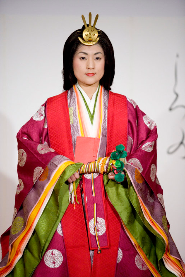 Eine Frau in einem traditionellen japanischen Kimono, das ein leuchtend rotes und grünes Kleid mit aufwändigen Mustern zeigt, steht selbstbewusst auf einem Laufsteg, ihr Haar ist ordentlich zu einem Knoten gebunden.