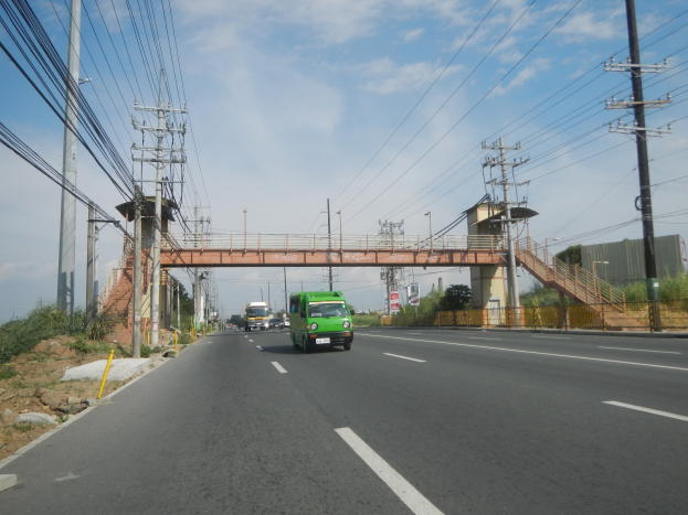 Grüner Lkw auf einer Straße neben einer Brücke, mit elektrischen Polen und Drähten entlang der Straße, Bäumen und Gebäuden im Hintergrund unter einem klaren blauen Himmel.