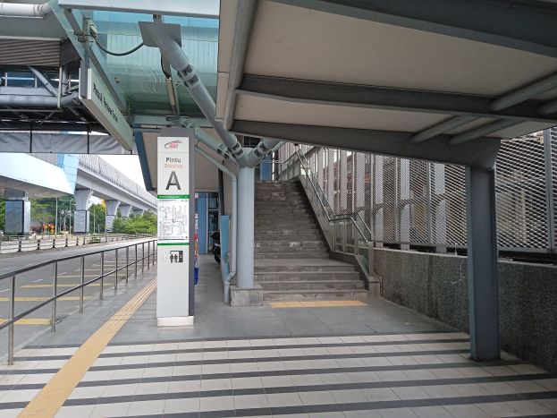 Ein Bahnsteig mit Treppe, eine Straße mit Geländer auf der linken Seite und Hintergrund-Elemente wie Bäume, Pfosten, Gebäude und eine Brücke; an der Spitze des Bahnsteigs sind Rohre und ein Schild zu sehen.