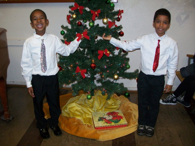 Ein geschmückter Weihnachtsbaum steht in einem Raum mit einem hölzernen Tisch auf der linken Seite und einem Stuhl auf der rechten Seite, zwei Jungen in weißen Hemden, die strahlen und den Baum halten, eine Schachtel und andere Gegenstände auf dem Boden und eine weiße Wand mit angebrachten Gegenständen im Hintergrund.