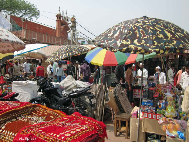 Eine belebte Szene mit Schirmen, Fahrzeugen, Matten, Kisten, einem Hocker, Menschen und verschiedenen anderen Gegenständen, mit einem Mikrofon, einer aktuellen Stange, Kabeln und einer Moschee im Hintergrund.