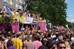 Eine große Menschenmenge marschiert auf der Straße bei Berlin Pride 2019, viele tragen Mützen und Schutzbrillen, halten Fahnen, mit Bannern und Gebäuden im Hintergrund bei bewölktem Himmel.