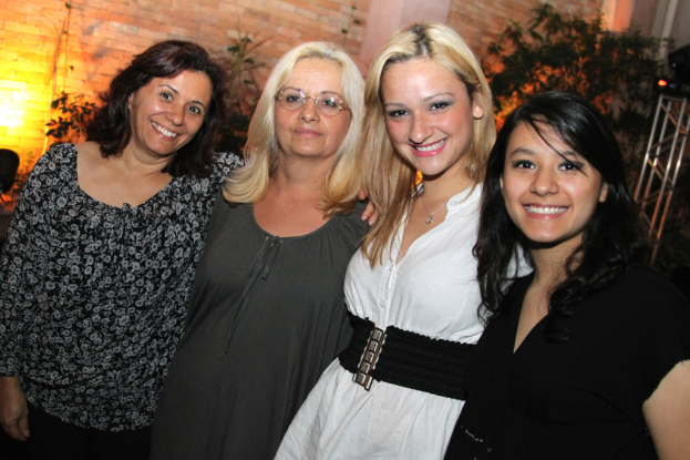 Vier Frauen in der Mitte, die in die Kamera löcheln und posen, mit einer Ziegelwand und Pflanzen im Hintergrund.