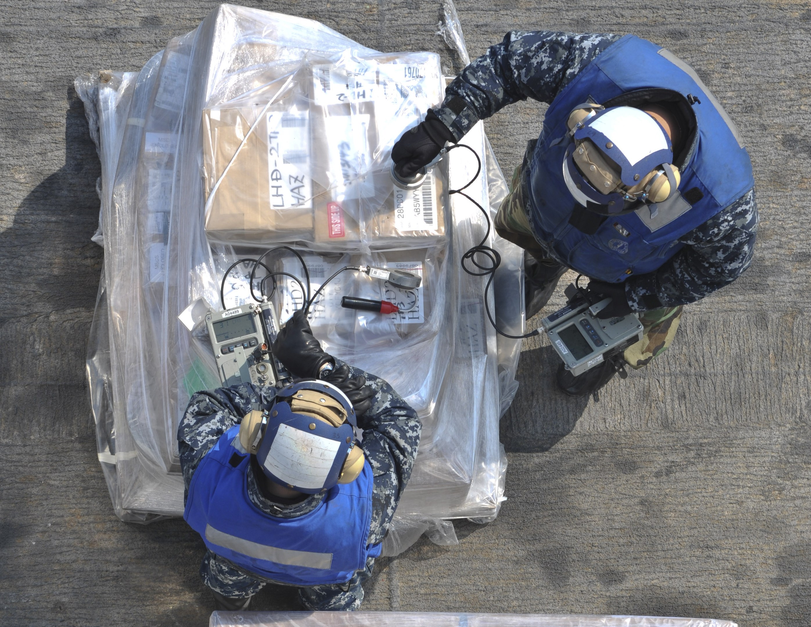 Zwei Männer in blauen Uniformen und Helmen arbeiten an einer großen Drohne, halten verschiedene Gegenstände während eine Plastikabdeckung auf dem Boden vor ihnen liegt.