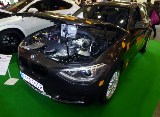 Ein BMW-Auto auf einer Autauschau mit offenem Motorhaube, umgeben von Menschen, mit einem Informationsschild auf einem Stand links und Säulen und einer Wand im Hintergrund.