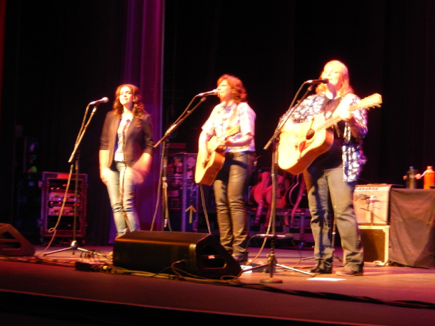 Drei Frauen treten auf der Bühne eines Musikkonzerts auf, jede singt in ein Mikrofon, während sie eine Gitarre spielt, mit einem Tisch voller Wasserflaschen und zusätzlichen Gitarren dahinter.