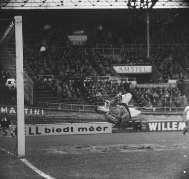 Ein Fußballer in Schwarz-Weiß-Fotografie hält vor einem Tor, mit Netz links und Zuschauern im Hintergrund sowie Texttafeln.