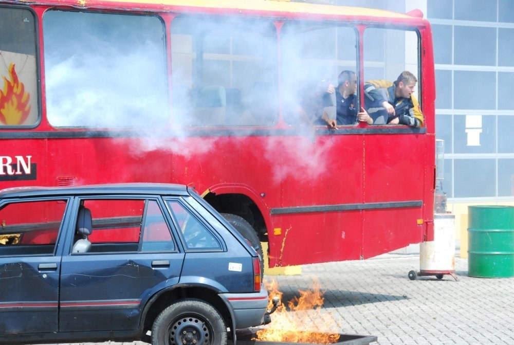Roter Doppeldeckerbus mit Rauch, daneben geparktes Auto, drei sichtbare Insassen und ein Gebäude mit Glasfenstern und einem Fass im Hintergrund.
