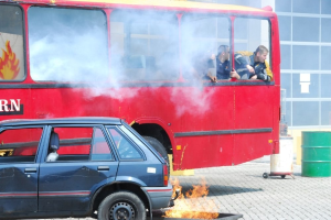 Roter Doppeldeckerbus mit Rauch, daneben geparktes Auto, drei sichtbare Insassen und ein Gebäude mit Glasfenstern und einem Fass im Hintergrund.