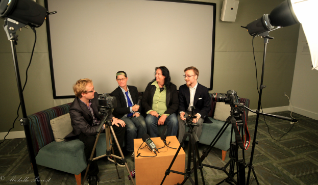 Eine Gruppe von drei Personen sitzt auf einem Sofa und gibt ein Interview, wobei eine Person links in einem schwarzen Mantel und Hose, eine Person in der Mitte in einem Mantel und blauer Krawatte mit einem Mikrofon und eine Person rechts mit einem strahlenden Lächeln zu sehen ist, sowie ein Projektionsbildschirm dahinter, ein Mikrofonrekorder auf einem Tisch in der Mitte und Kameras mit großen Stativen auf beiden Seiten.