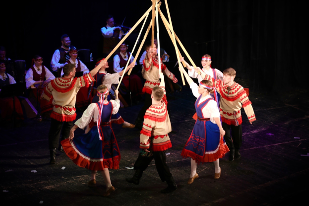 Eine Gruppe von Menschen in farbenfrohen Kostümen tanzt um einen Maibaum, einige halten Holzstöcke, während andere im Hintergrund Musikinstrumente spielen.