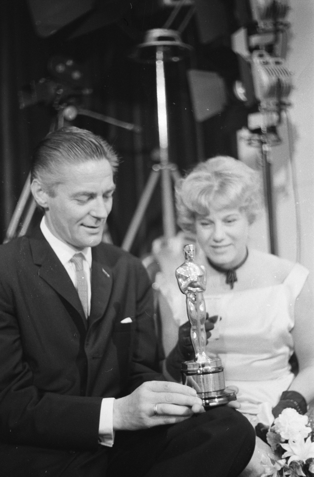 Ein Mann in einem schwarzen Blazer, weißem Hemd und Krawatte hält eine Oscar-Statue, während er neben einer Frau in Abendgarderobe sitzt, die einen Blumenstrauß hält, mit weicher Beleuchtung im Hintergrund.