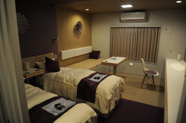 Hotelzimmer mit zwei Betten, jeweils mit Decken und Kissen, einem Schreibtisch mit Gegenständen, einem Stuhl, einem Vorhang, einer Klimaanlage, Deckenleuchten und an der Wand angebrachten Gegenständen.