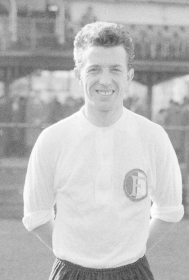 Ein Schwarz-Weiß-Foto eines grinsenden Mannes in einem T-Shirt und Shorts auf einem Fußballfeld, mit einem Gebäude im Hintergrund, das mutmaßlich Mario Mandžukić ist.
