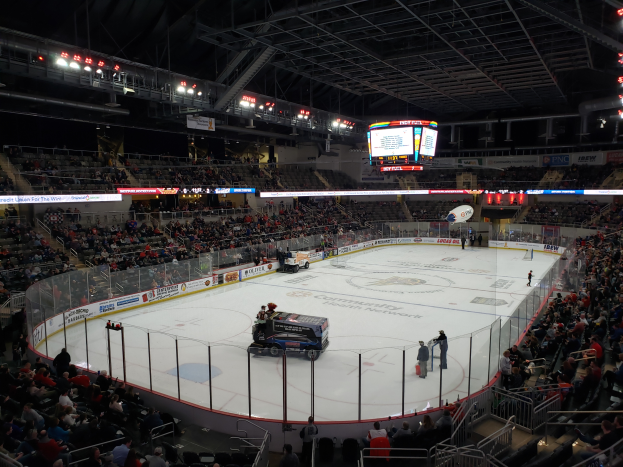 Ein Eishockeyspiel in einer großen Arena mit Fahrzeugen auf dem Eis, Zuschauern rund um die Bahn und Menschen auf den Tribünen, sowie Boards, Lichter und ein Display im Hintergrund.