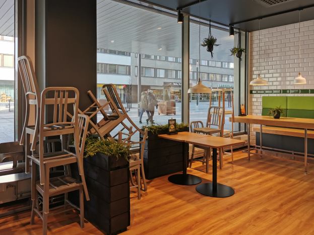 Ein Restaurant in Berlin mit Holztischen und -stühlen, umgeben von Pflanzen und Lichtern, mit einem großen Fensterblick auf Menschen auf der Straße, Gebäude und den Himmel.