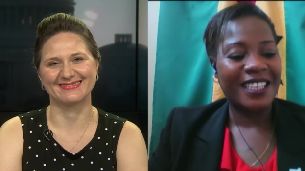 Zwei Frauen, die während eines Videoanrufs lächeln, eine in einem schwarzen und weißen Kleid, die andere in einer unbestimmten Farbe, mit einer Flagge, die an der Wand hinter ihnen in einer öffentlichen Einrichtung hängt.