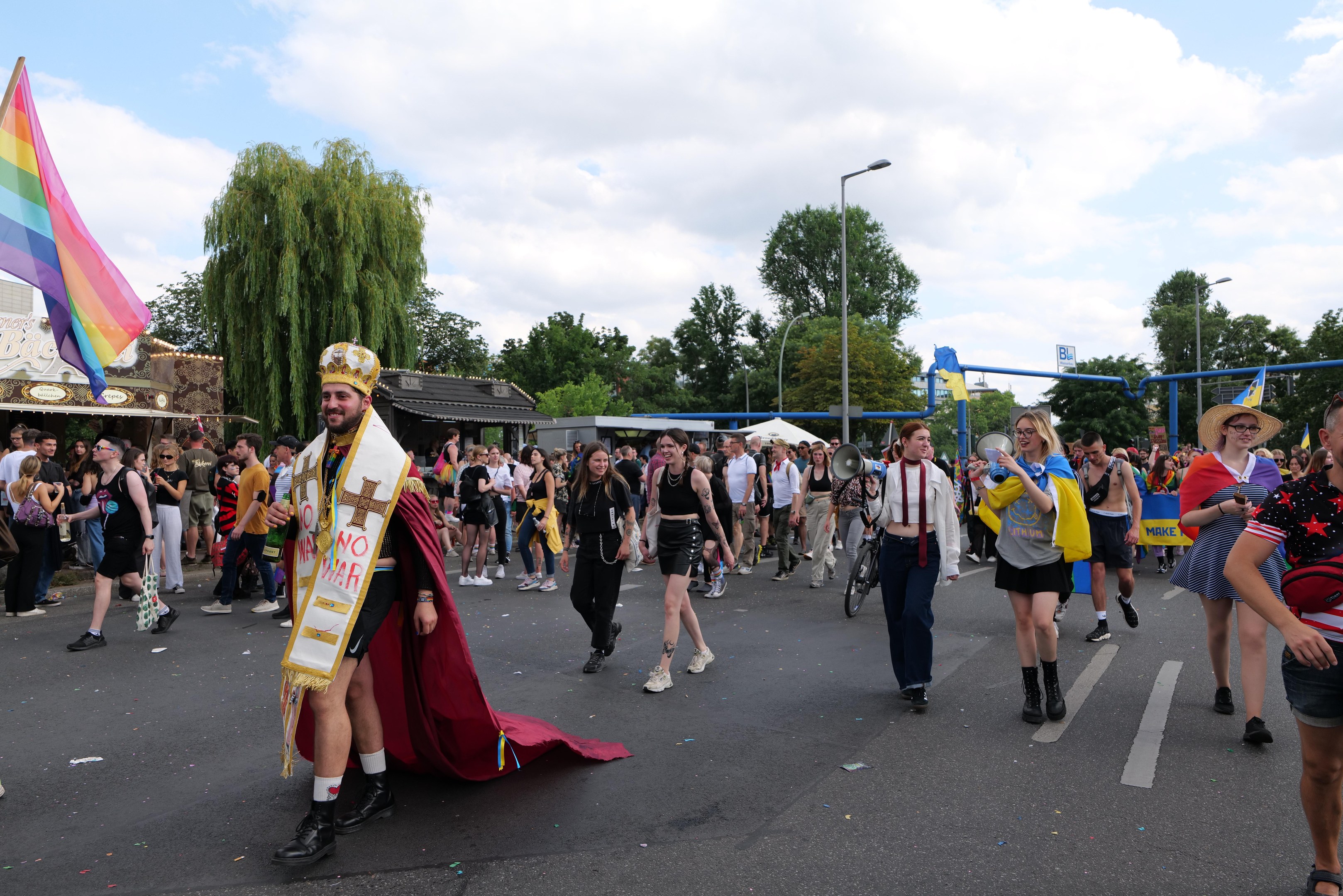 Eine Gruppe von Menschen marschiert bei der Pride Parade 2018 mit einer Regenbogenfahne und Musikinstrumenten, im Hintergrund sind Laternenpfähle, Bäume, Hütten und ein bewölkter Himmel zu sehen.