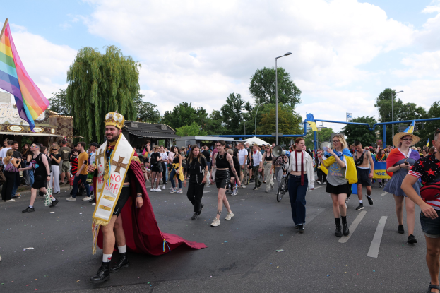 Eine Gruppe von Menschen marschiert bei der Pride Parade 2018 mit einer Regenbogenfahne und Musikinstrumenten, im Hintergrund sind Laternenpfähle, Bäume, Hütten und ein bewölkter Himmel zu sehen.