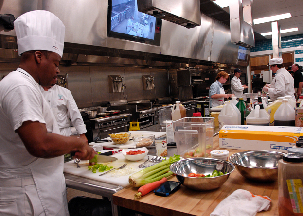 Eine Gruppe von Köchen in einer Küche, die Essen auf einem Schneidbrett zubereiten, mit einem Koch im Vordergrund in einer weißen Schürze und Mütze, der ein Messer hält, und umgeben von verschiedenen Utensilien, Schüsseln, Gemüse, einem Mobiltelefon, Flaschen und anderen Gegenständen, mit einem Fernseher und Deckenlampen im Hintergrund.
