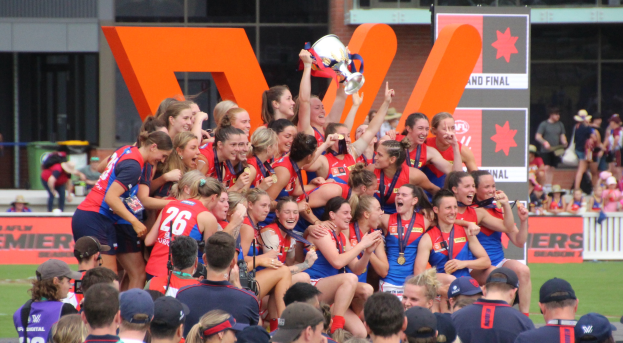 Eine Gruppe von Frauen-AFL-Spielerinnen in Sportuniformen feiert mit einem Pokal vor einem Publikum, einige halten ihn, während andere daneben stehen.